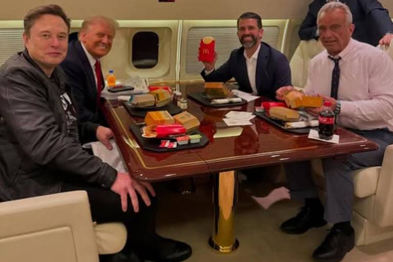 Cinco homens estão sentados em uma mesa dentro de um jato particular， com comida de fast food à sua frente. Um deles está segurando uma batata frita da McDonald039;s. O ambiente é luxuoso， com assentos em couro e uma mesa de madeira polida. Todos parecem estar se divertindo.