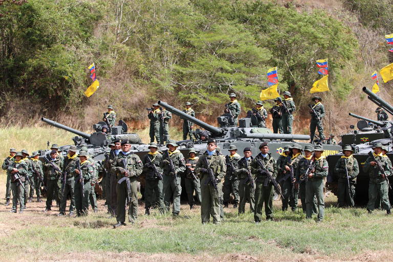 A imagem mostra um grupo de soldados em um campo， com tanques de guerra ao fundo. Os soldados estão vestidos com uniformes militares e alguns estão armados. Bandeiras com cores da Venezuela estão visíveis， e a vegetação ao redor é verde e montanhosa.