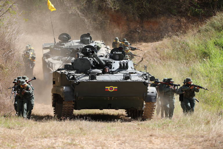 A imagem mostra um cenário de operação militar em um terreno aberto. Dois veículos blindados estão em destaque, com um deles à frente e outro um pouco atrás. Soldados armados estão se movendo em direção ao espectador, alguns agachados e outros em pé, todos usando uniformes camuflados. Ao fundo, há vegetação e poeira levantada, indicando movimento. Uma bandeira amarela está visível em um dos veículos.