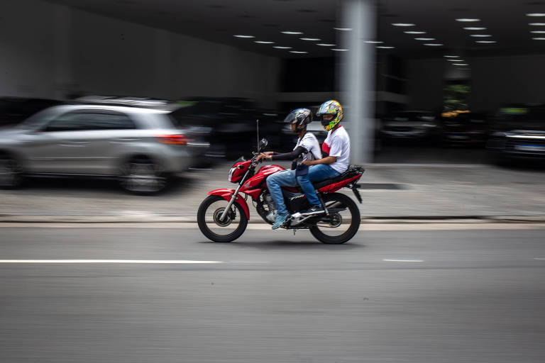 A imagem mostra uma motocicleta vermelha em movimento, com duas pessoas a bordo. O motociclista está usando um capacete amarelo e a pessoa na garupa está vestindo uma camiseta branca. Ao fundo, há uma calçada e um carro estacionado