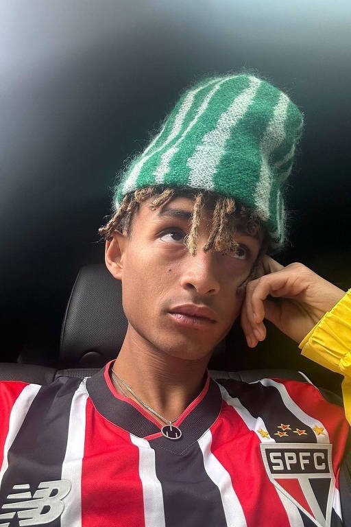 A imagem mostra um jovem sentado em um carro, usando uma camiseta de futebol com listras vermelhas e brancas, que representa o São Paulo FC. Ele está usando um gorro verde com listras brancas e tem cabelo cacheado. O jovem está olhando para cima, com a mão apoiada no rosto, e usa um colar. A luz do ambiente é suave, criando um efeito de iluminação interessante.