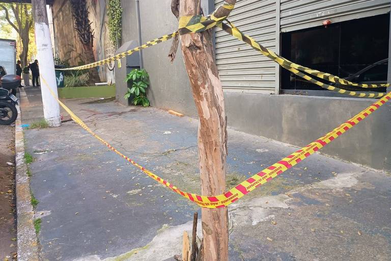 A imagem mostra um trecho de calçada que está bloqueado por fitas de segurança amarelas e pretas， amarradas em um tronco de árvore e em uma estrutura próxima. Ao fundo， há uma parede com janelas e uma planta visível. O ambiente parece urbano.