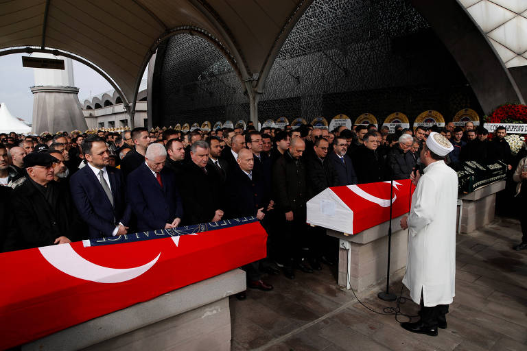 A imagem mostra uma cerimônia de homenagem com várias pessoas reunidas em um espaço coberto. No centro， há dois caixões cobertos com bandeiras da Turquia. À frente， um homem vestido de branco， possivelmente um líder religioso， está realizando uma cerimônia. Os presentes estão em pé， com expressões sérias， demonstrando respeito pela ocasião