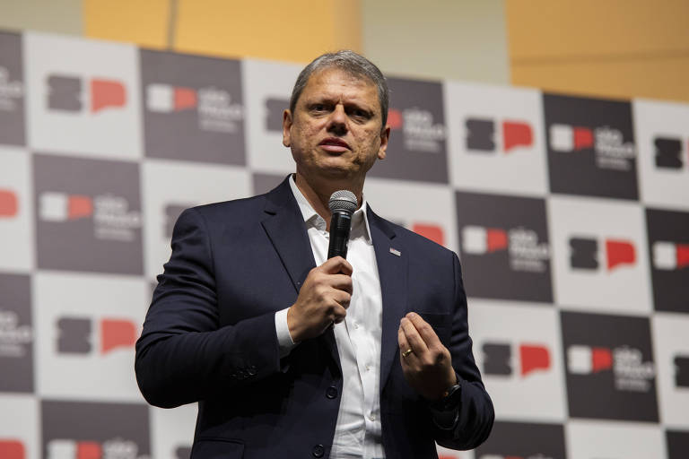 Um homem em um terno escuro está falando ao microfone em um evento. Ele parece estar explicando algo， gesticulando com a mão. Ao fundo， há um painel com um padrão de quadrados em preto e branco. É Tarcísio de Freitas， governador de São Paulo