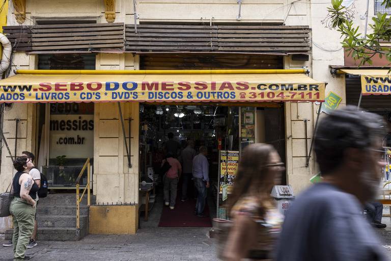 A imagem mostra a fachada de uma loja chamada 'Sebo do Messias', que vende livros, CDs, DVDs e discos. O letreiro é amarelo e azul, com o endereço do site e o número de telefone visíveis. Algumas pessoas estão passando pela calçada em frente à loja, e a entrada da loja é visível, com uma escada que leva ao interior.