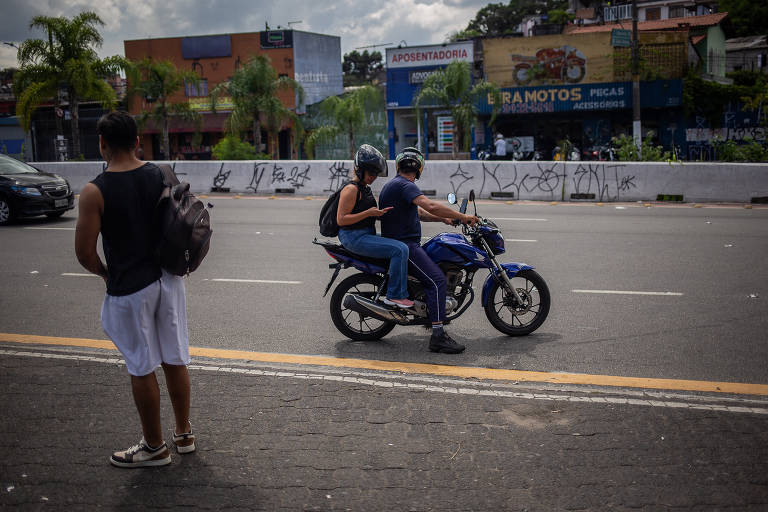 A imagem mostra uma motocicleta azul, com duas pessoas a bordo, no meio de uma avenisa. Um homem está parado na calçada, observando a cena. Ao fundo, há prédios e outdoors visíveis, além de algumas palmeiras. O céu está parcialmente nublado.
