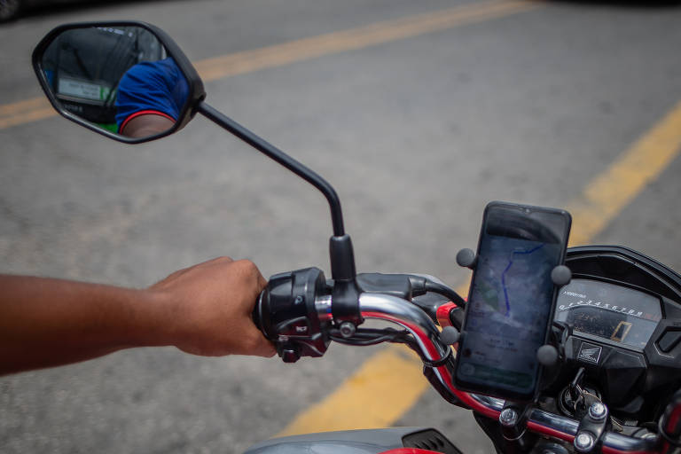 A imagem mostra uma mão segurando o guidão de uma motocicleta. O guidão é prateado e possui um espelho retrovisor à esquerda， refletindo parte do ambiente. Um celular está preso em um suporte no guidão， visível na parte frontal. O fundo apresenta uma estrada com faixas amarelas.
