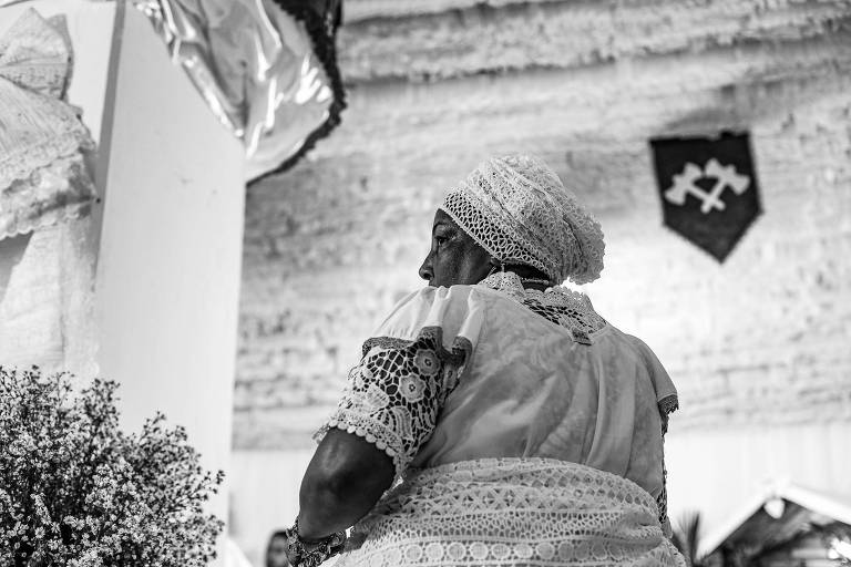 A imagem em preto e branco mostra uma mulher de costas， vestindo um traje tradicional branco com detalhes em renda. Ela usa um lenço na cabeça e está em um ambiente decorado， com um fundo que apresenta uma parede de pedra e um brasão com dois machados cruzados. Há também um arranjo de flores na parte inferior esquerda da imagem.