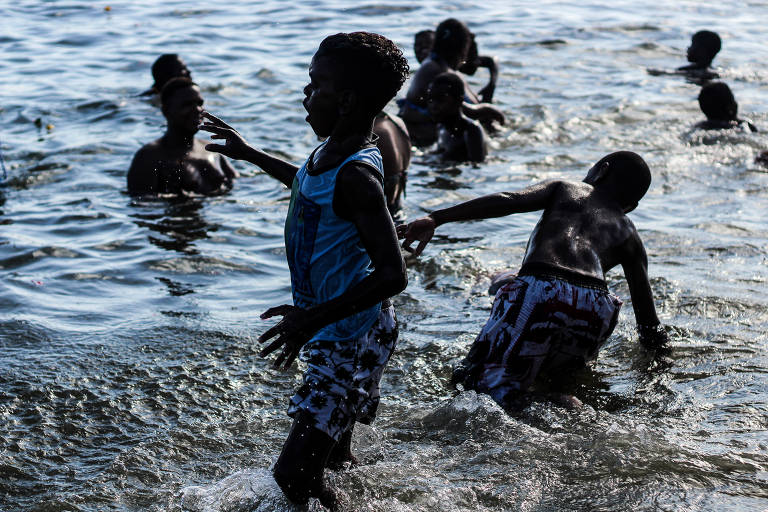 A imagem mostra várias crianças brincando na água. Um menino em primeiro plano, com uma camiseta azul e shorts estampados, está correndo em direção à câmera, enquanto outros meninos nadam e brincam ao fundo. A água está agitada, refletindo a luz do sol, e a cena transmite uma sensação de diversão e alegria.