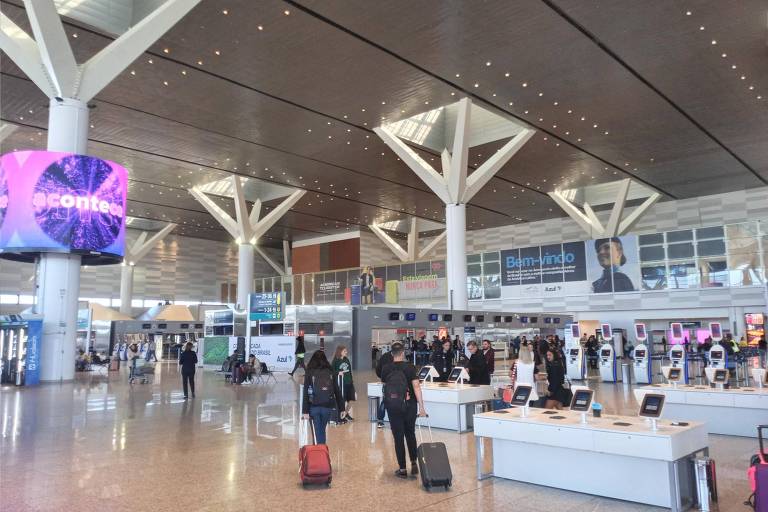 A imagem mostra o interior de um aeroporto moderno， com um grande espaço aberto e tetos altos. Há várias pessoas se movendo com malas， algumas paradas em balcões de check-in. O piso é de cerâmica clara e há colunas estruturais visíveis. Ao fundo， há um grande painel digital com cores vibrantes.