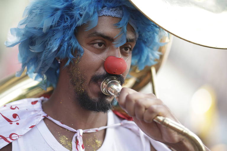 Um palhaço com cabelo azul e nariz vermelho está tocando um instrumento de metal. Ele usa uma camisa branca com detalhes coloridos e parece estar em um ambiente festivo.