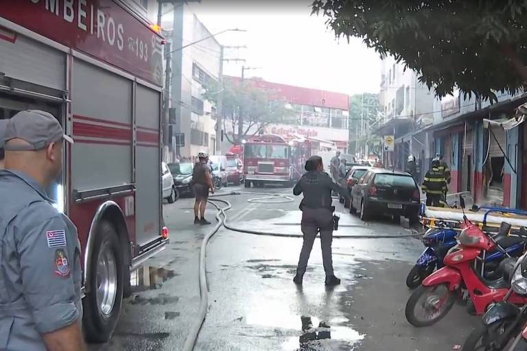 A imagem mostra uma cena de incêndio em uma área urbana. Há um caminhão de bombeiros vermelho à esquerda e várias pessoas， incluindo um bombeiro com mangueira， tentando controlar as chamas. O ambiente está cheio de fumaça e há veículos estacionados ao longo da calçada， incluindo várias motos vermelhas. O cenário parece caótico devido à situação de emergência.