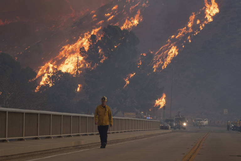 A imagem mostra um incêndio florestal em uma encosta montanhosa， com chamas intensas subindo entre as árvores. Um bombeiro， vestido com uniforme de combate a incêndios e capacete， caminha em direção à câmera， enquanto veículos de emergência estão estacionados ao lado da estrada. O céu está coberto de fumaça， criando uma atmosfera densa e sombria.