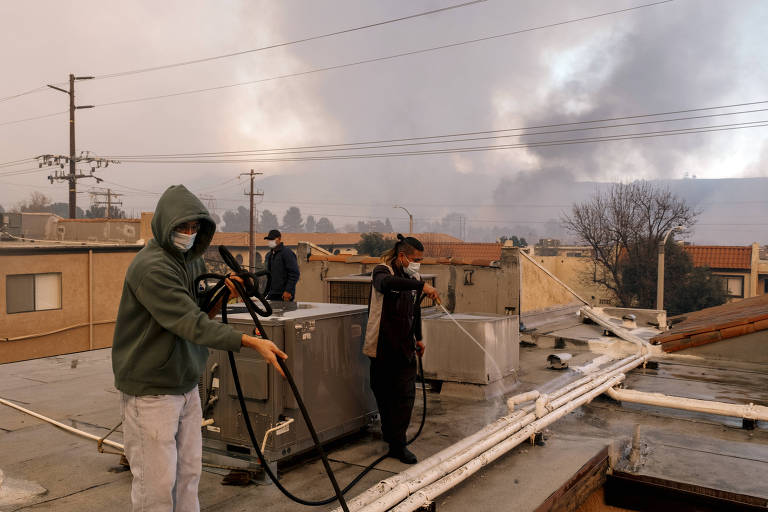 A imagem mostra três pessoas em um telhado， trabalhando em meio a uma situação de incêndio. Um homem à esquerda， usando um moletom verde e máscara， está manuseando uma mangueira. Outro homem， ao centro， também com máscara， está usando uma mangueira para limpar. Ao fundo， uma terceira pessoa observa. O céu está nublado e há fumaça visível， indicando a presença de um incêndio nas proximidades.