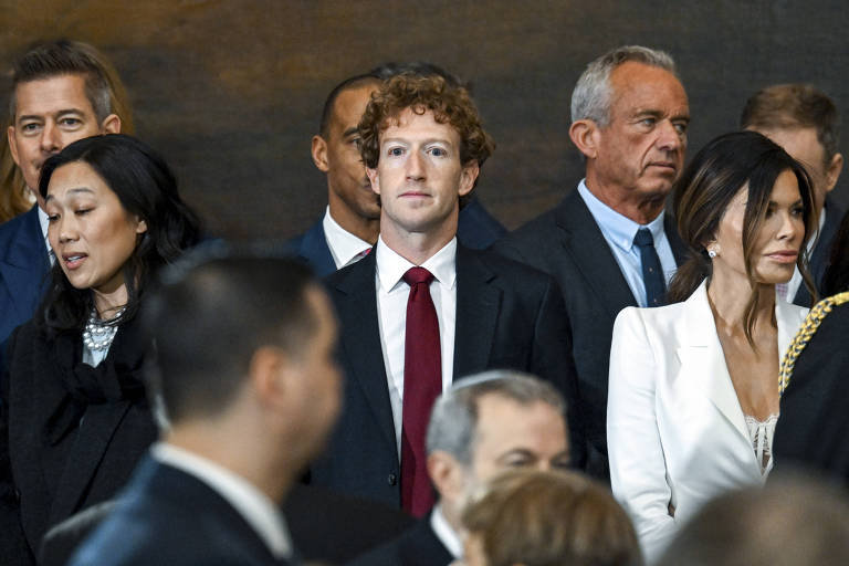 A imagem mostra um grupo de pessoas em um evento formal. No centro， um homem com cabelo cacheado e terno escuro， usando uma gravata vermelha. À esquerda， uma mulher com cabelo longo e escuro， e à direita， uma mulher vestindo um vestido branco. Ao fundo， outras pessoas também estão presentes， algumas com expressões sérias.
