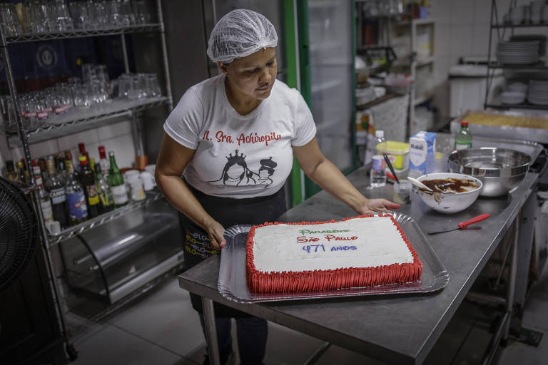 Uma confeiteira está decorando um bolo retangular em uma cozinha. Ela usa uma camiseta branca e um touca de proteção. O bolo tem cobertura branca com detalhes em vermelho e uma mensagem escrita em cima， que diz quot;parabéns， são paulo， 471 anosquot;. Ao fundo， há prateleiras com utensílios de cozinha e bebidas.