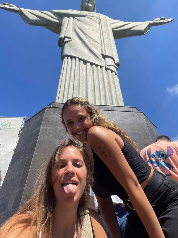 Duas jovens brancas e loiras posam na frente da estátua do Cristo Redentor no Rio. Uma está de língua de fora， a outra， sorrindo