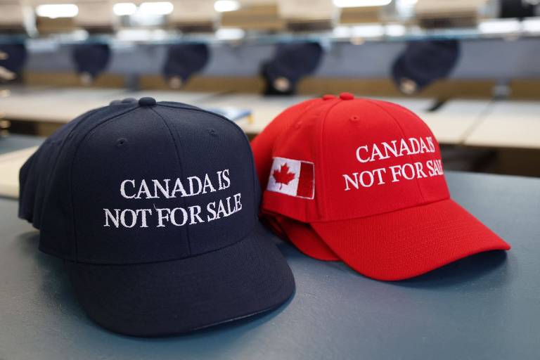 A imagem mostra dois bonés sobre uma superfície. O boné à esquerda é azul escuro e possui a frase 039;CANADA IS NOT FOR SALE039; em letras brancas. O boné à direita é vermelho e também apresenta a mesma frase， além de uma pequena bandeira do Canadá na lateral. Ao fundo， há uma mesa com outros objetos， mas não estão claramente visíveis.
