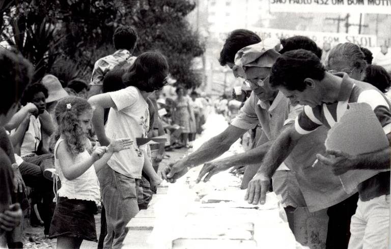 Foto em preto e branco mostra pessoas manuseando um longo bolo, e elas são observadas por um grande público