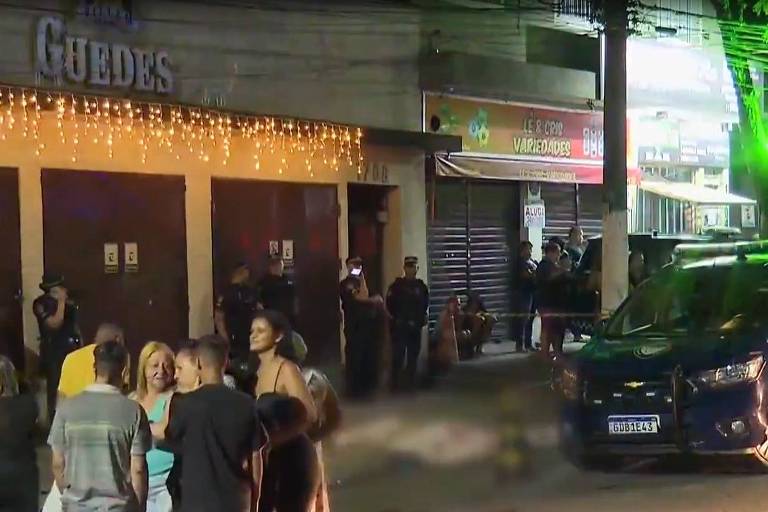 A imagem mostra uma cena noturna em frente ao estabelecimento chamado 039;Guedes039;. Há várias pessoas reunidas do lado de fora， algumas conversando e outras observando. Vários policiais estão presentes， alguns em pé e outros em uma viatura da Guarda Civil. O local está decorado com luzes penduradas. 