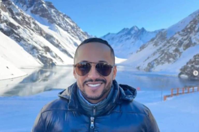 Um homem sorridente está posando em frente a uma paisagem montanhosa coberta de neve. Ele usa óculos escuros e uma jaqueta escura. Ao fundo， há montanhas nevadas e um lago refletindo o céu azul claro.