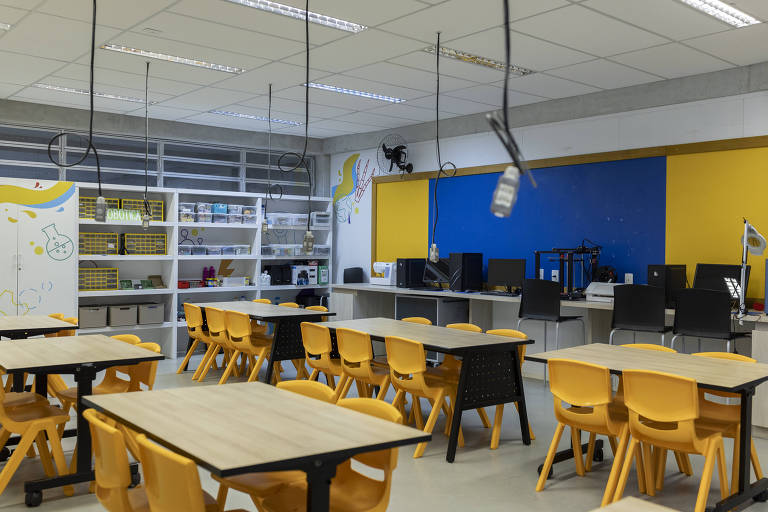 A imagem mostra uma sala de aula moderna com mesas e cadeiras amarelas dispostas em um arranjo organizado. Ao fundo， há uma parede azul e amarela， além de prateleiras com materiais. Equipamentos de informática estão visíveis em mesas， e a iluminação é clara e uniforme.
