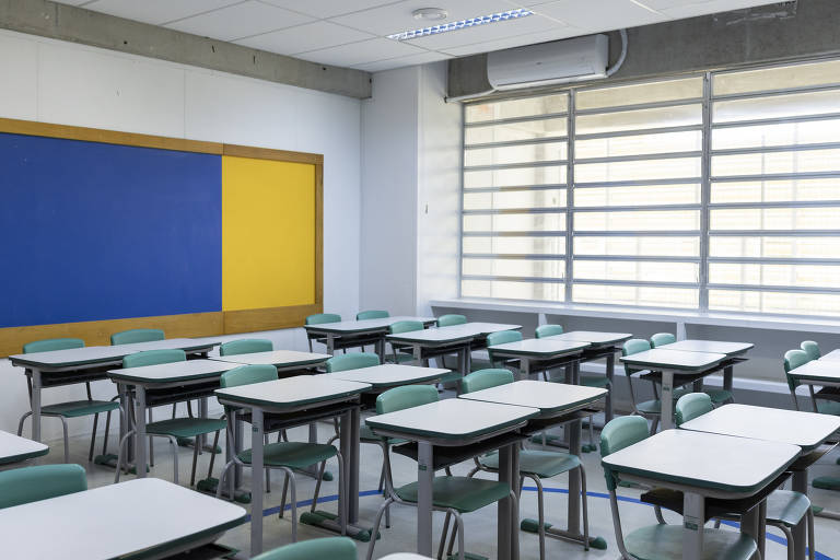 A imagem mostra uma sala de aula com mesas e cadeiras organizadas. As mesas são brancas com pernas pretas e as cadeiras são verdes. Há uma parede com um quadro azul e uma parte amarela. Uma janela com persianas permite a entrada de luz natural.