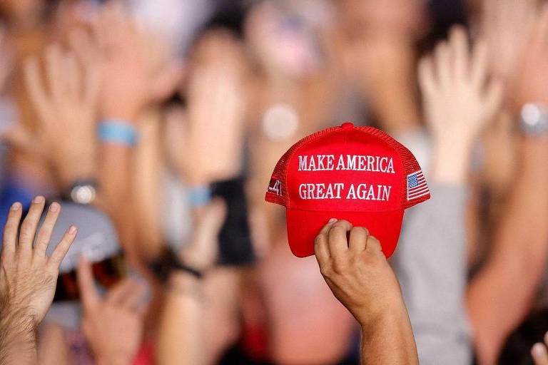 A imagem mostra um chapéu vermelho com a frase 039;MAKE AMERICA GREAT AGAIN039; em destaque， segurado por uma mão. Ao fundo， várias mãos levantadas de pessoas em uma multidão， sugerindo um evento ou comício.
