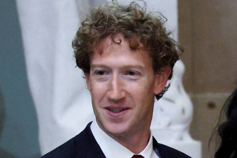 A imagem mostra Mark Zuckerberg， um homem com cabelo cacheado e claro， usando um terno escuro. Ele está sorrindo e parece estar em um ambiente interno， com esculturas ou estátuas ao fundo.