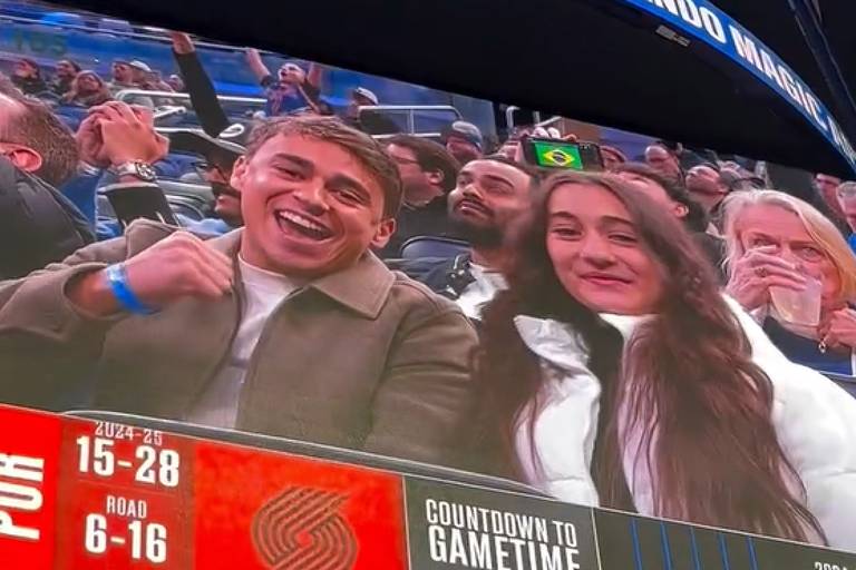 A imagem mostra um telão em um evento esportivo, onde um jovem e uma jovem estão sorrindo e posando para a câmera. O telão exibe as palavras 'BLAZERS vs MAGIC' e informações sobre o jogo, incluindo contagem regressiva para o início e estatísticas das equipes. Ao fundo, há outros torcedores assistindo ao jogo.