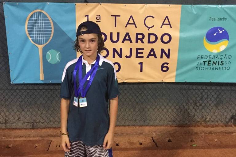 Um jovem atleta está posando em frente a um banner que anuncia a 1ª Taça Eduardo Bonjean, realizada em 2016. Ele usa uma camiseta verde escura com detalhes brancos e calções listrados em azul e branco. O atleta tem duas medalhas penduradas no pescoço e um boné. O fundo do banner apresenta uma raquete de tênis e bolas, além do logotipo da Federação de Tênis do Rio de Janeiro.