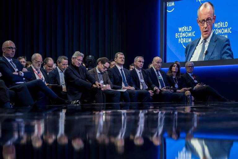 A imagem mostra um painel de discussão em um evento， com várias pessoas sentadas em fileira， prestando atenção a um orador que aparece em um grande telão ao fundo. O orador está usando óculos e parece estar falando sobre um assunto importante. O ambiente é escuro， com iluminação focada no painel e no telão.