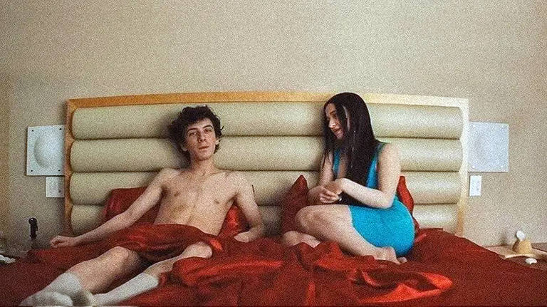  A imagem mostra um jovem homem sem camisa e uma jovem mulher vestindo um vestido azul， sentados em uma cama com lençóis vermelhos. O ambiente é um quarto com paredes claras e um cabeceira de cama em tom claro. O homem parece pensativo， enquanto a mulher sorri e parece estar conversando com ele.