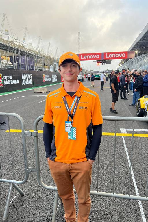 Um jovem está posando em um evento automobilístico, vestindo uma camisa e um boné laranja com o logotipo da McLaren. Ele está em uma área delimitada por grades, com o fundo mostrando uma pista de corrida e várias pessoas ao redor. Ao fundo, há uma placa com os dizeres 'CITY OF SÃO PAULO' e 'ALWAYS AHEAD', além de um sinal de 'Lenovo'. O céu está nublado.