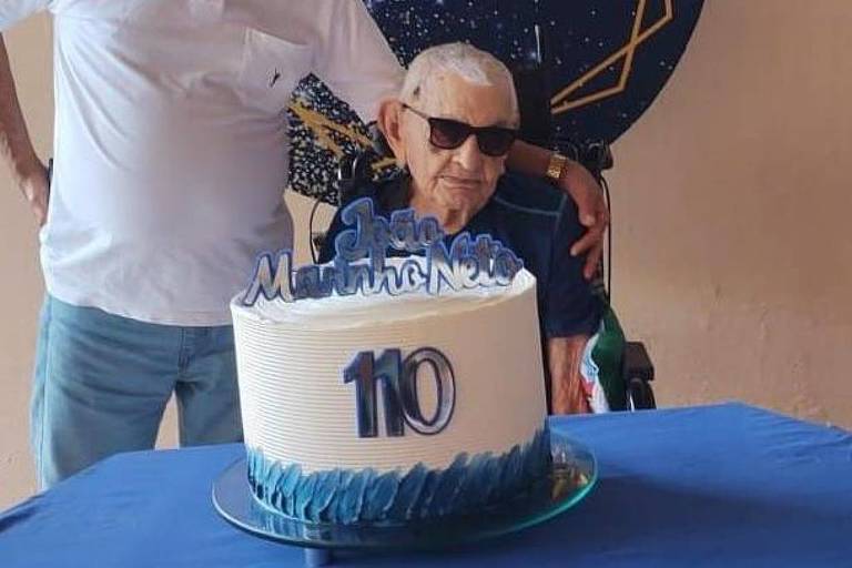 Um homem idoso， usando óculos escuros， está sentado em uma cadeira ao lado de um bolo de aniversário. O bolo é branco com detalhes em azul e tem o número 039;110039; em destaque. Ao fundo， uma pessoa está parcialmente visível， com uma camisa branca. A mesa é coberta com uma toalha azul.