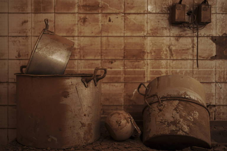 A imagem mostra uma cozinha abandonada com panelas de metal sujas e desgastadas. Uma panela está virada， enquanto outra está de pé ao lado de uma panela maior. No chão， há um objeto que parece ser uma cabeça de boneco. As paredes estão cobertas de azulejos sujos e manchados， com marcas de umidade e sujeira visíveis.