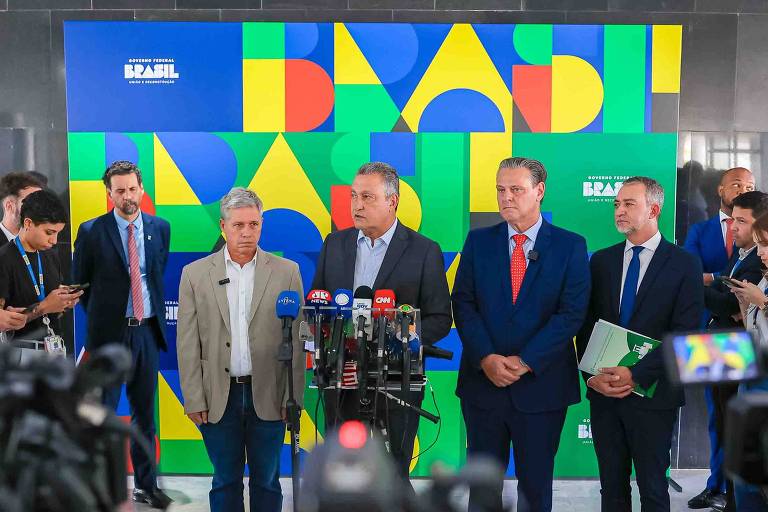 A imagem mostra uma coletiva de imprensa com várias pessoas em pé em frente a um fundo colorido que diz 039;BRASIL039;. No centro， um homem fala ao microfone， cercado por outros quatro homens. Há jornalistas e câmeras ao redor， capturando o evento.