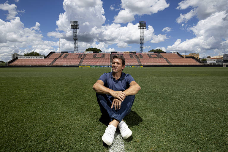 Juninho Paulista no gramado do Estádio Novelli Júnior， em Itu. Um homem está sentado no gramado de um campo de futebol， com as pernas cruzadas e uma expressão pensativa. Ele usa uma camiseta azul e calças jeans， além de tênis brancos. Ao fundo， há uma arquibancada vazia e torres de iluminação sob um céu com nuvens brancas e azuis.
