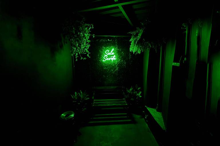 A imagem mostra um espaço com iluminação verde, onde uma parede com plantas é iluminada por um letreiro neon que diz 'Sala Secreta'. Há um banco de madeira escura e algumas plantas ao redor, além de um recipiente de lixo visível à esquerda.