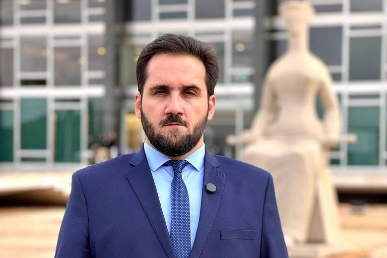 Homem com barba rala e bigode， cabelo escuro， de terno azul marinho， com camisa azul claro e gravata azul， fotografado da cintura para cima， de pé， em frente à estátua da Justiça que fica na área externa do Supremo Tribunal Federa， em Brasília