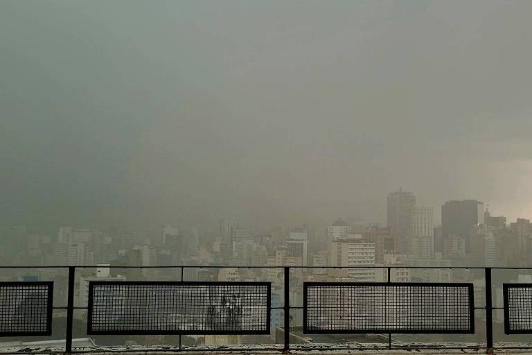 A imagem mostra uma vista panorâmica de parte da cidade com nuvens escuras cobrindo o céu. No primeiro plano， há uma grade de proteção， e ao fundo， é possível ver vários edifícios e arranha-céus. 