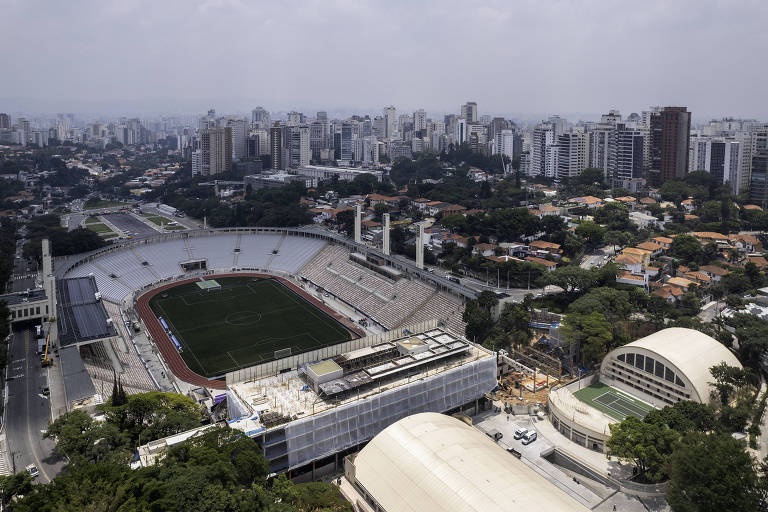 Imagem aérea de um estádio de futebol com campo verde e arquibancadas vazias， cercado por áreas urbanas e prédios altos. À direita， há uma estrutura arquitetônica de formato curvo e outras construções. O céu está nublado， e a vegetação é visível ao redor do estádio.