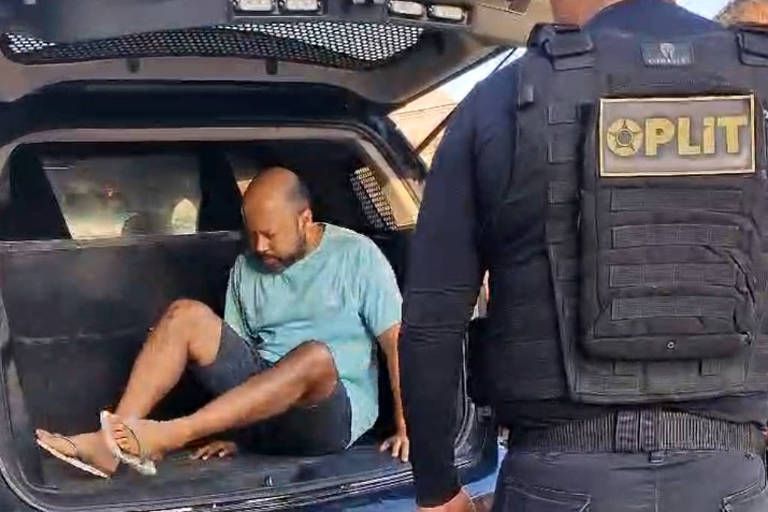 Imagem mostra o serial killer de camisa azul dentro da viatura da polícia， sendo acompanhado por um agente. 