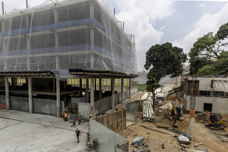 A imagem mostra um edifício em construção， parcialmente coberto por uma lona. O prédio possui várias andares e está cercado por uma área de obras， onde há máquinas e trabalhadores. Ao fundo， é possível ver uma árvore e outras estruturas em construção