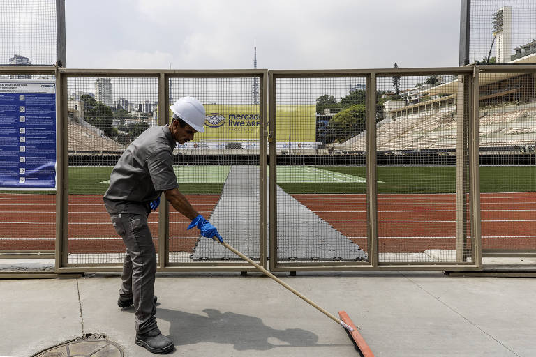 Um trabalhador está usando um rodo para limpar o chão de um estádio, em frente à grade de um alambrado. Ele está vestido com uma camisa cinza e calças escuras, usando luvas azuis e um capacete branco. Atrás da grade, há uma pista de atletismo de cor laranja; A fundo, é possível ver as arquibancadas do estádio e uma faixa amarela com o texto 'Mercado Pago Arena'.