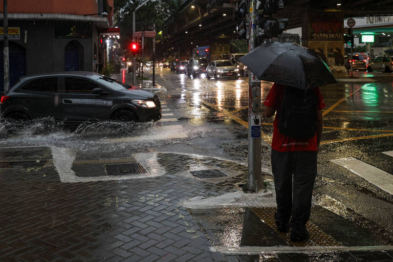 A imagem mostra uma cena urbana durante uma forte chuva. Um homem está de pé em uma calçada， segurando um guarda-chuva preto. Ele está vestido com uma camiseta vermelha e calças escuras. Ao fundo， um carro cinza passa por uma poça de água， levantando respingos. As ruas estão molhadas e há luzes de trânsito visíveis， além de outros veículos se movendo. O ambiente parece movimentado， mesmo sob a chuva.