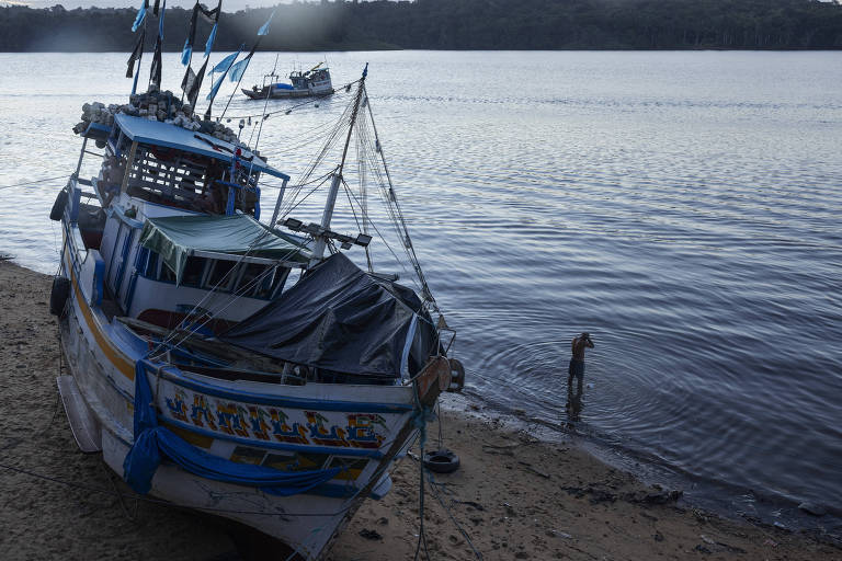 A imagem mostra um barco de pesca ancorado na margem de um rio， com bandeiras azuis e brancas. À direita， uma pessoa está em pé na água， enquanto o barco está parcialmente coberto por uma lona. O ambiente é tranquilo， com a superfície da água refletindo a luz do dia.
