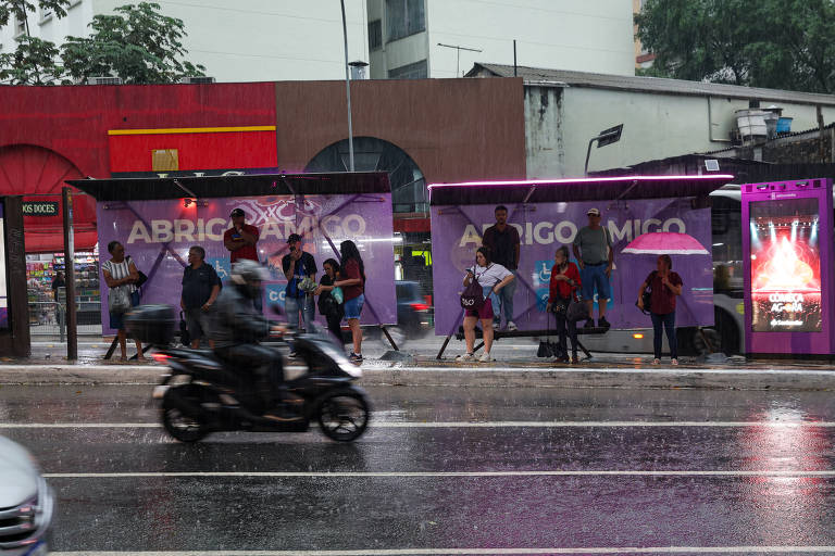 A imagem mostra um ponto de ônibus durante uma chuva intensa. Algumas pessoas estão esperando o ônibus sob a cobertura， enquanto outras estão se protegendo da chuva com guarda-chuvas. Um motociclista passa pela rua alagada. O cenário é urbano， com prédios ao fundo e uma iluminação suave.