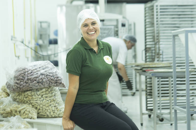 Na foto， Maíria Welerson está sentada em uma mesa， usando uma touca. Ela posa em área de produção， exibindo produtos da empresa