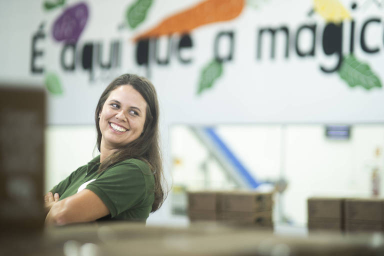 Na foto， Maíra Welerson， mulher branca， de cabelo castanho comprido， sorri olhando para o lado. Ela usa camisa polo verde-musgo de manga curta e está com os braços cruzados. Ao fundo há um grande letreiro com a frase quot;É aqui que a mágica começaquot; parcialmente legível.